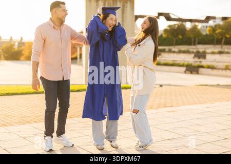 Three friends celebrate graduation day outdoors. The graduate, wearing a blue cap and gown, is joyfully embraced by two friends, enjoying a moment of Stock Photo