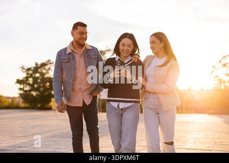 A group of three friends strolling outdoors, enjoying each other's company while sharing a moment on a mobile device. The sun sets behind them, creati Stock Photo
