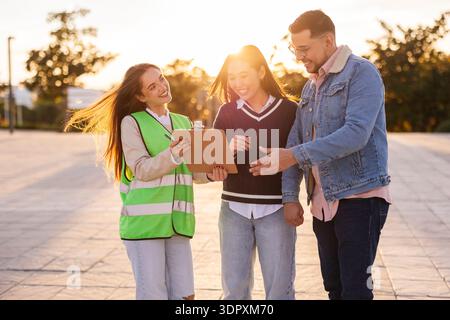 A group of diverse students stands outdoors, engaging in a friendly discussion. One holds a clipboard while the others listen attentively, basking in Stock Photo