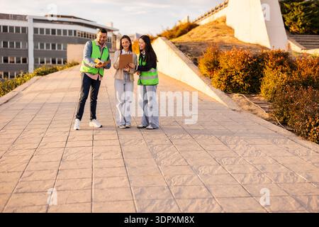 Three students wearing reflective vests discuss their project outdoors, using a clipboard and digital devices under sunny skies on a campus walkway. Stock Photo