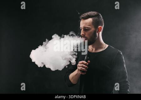 Man Exhales Large Cloud of Vapor While Using Vape Device in Dark Setting With Smoke in the Air Stock Photo