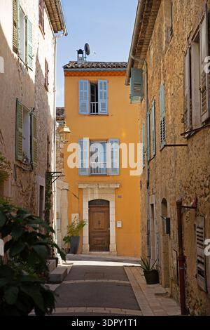 Narrow alley in Valbonne, Provence, with pastel shutters and a wooden door framed by warm ochre walls and stone facades. Stock Photo
