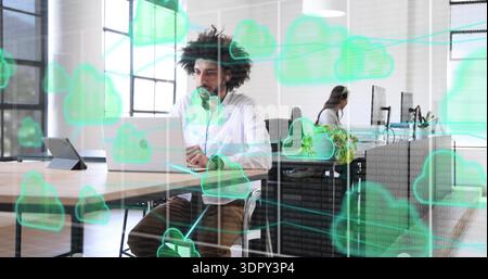 Working man in office wearing white shirt typing on laptop with headset and cloud overlays Stock Photo