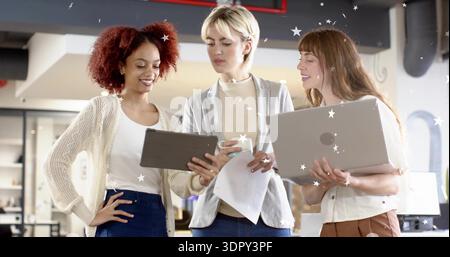 Reviewing three women colleagues wearing office attire, checking tablet and laptop in open office Stock Photo