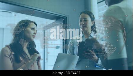 Presenting three women in blazers discussing digital map on office glass, holding tablet and laptop Stock Photo