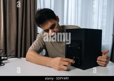 Focused young man inserting power cord into back of black studio monitor speaker, ensuring secure connection for audio setup on clean white table Stock Photo
