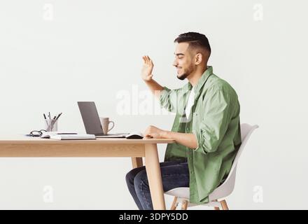 Man Waves During Video Call From Home Office Space With Laptop and Notebooks Stock Photo