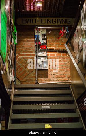 Stairway leading up to a brick-walled entrance with a "bad-eingang" sign, metal railings, dim industrial lighting, and posters along the walls. Stock Photo