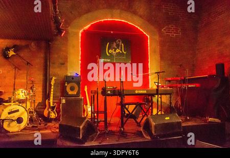 Empty live music stage in a cozy brick-arched venue with red neon backlight, drum kit, guitars, keyboard, amps, microphones and floor monitors, ready Stock Photo