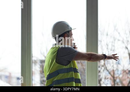 Middle aged Caucasian man wearing safety vest and hard hat standing in office, talking on walkie talkie, gesturing with arm extended toward window, overseeing construction project Stock Photo