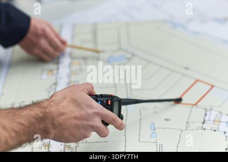 Middle aged Caucasian man holding walkie talkie and pointing pencil at architectural blueprint, working in office environment, demonstrating foreman responsibilities and coordination Stock Photo