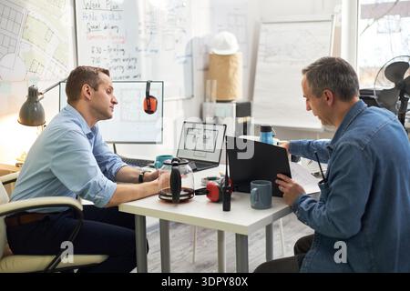 Middle aged Caucasian male engineer discussing construction project with foreman colleague in office, both reviewing architectural plans on laptop and printed documents at desk Stock Photo