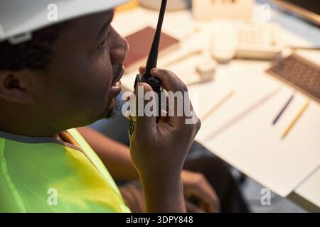Middle aged Black man wearing safety vest and hard hat using walkie talkie while sitting at office desk, communicating instructions, holding radio device near mouth, paperwork visible Stock Photo