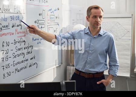 Middle aged Caucasian man standing in office pointing at whiteboard with construction schedule, presenting project plan to unseen audience, wearing business attire, serious expression Stock Photo
