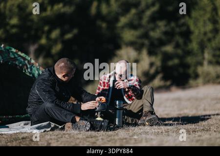 Two friends camping outdoors share a moment at a campsite with a stove and tent Stock Photo