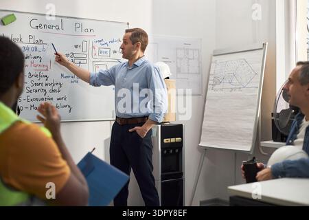 Caucasian middle aged man standing in office pointing at whiteboard with construction plans, while multiethnic group of building workers listening and taking notes during meeting Stock Photo