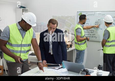 Middle aged Caucasian man collaborating with Black man in safety vest and helmet while reviewing construction plans in office, two young adult men in background discussing project at whiteboard Stock Photo