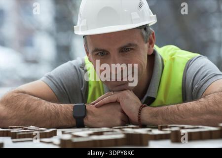 Middle aged Caucasian man wearing hard hat and safety vest leaning on table, studying architectural model pieces in office, foreman focusing on construction planning process Stock Photo