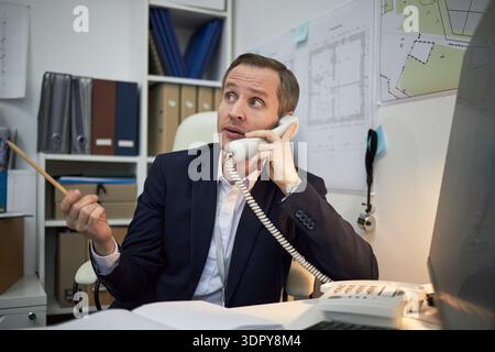 Middle aged Caucasian male engineer or architect sitting at office desk talking on landline phone, holding pencil, gesturing with hand, surrounded by architectural blueprints and documents Stock Photo