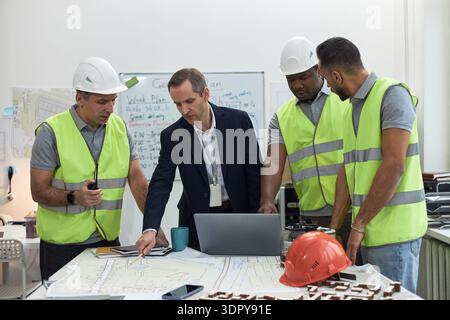 Middle aged Caucasian man in suit collaborating with two middle aged men and one young adult man in safety vests and helmets, discussing construction plans in office, using laptop and blueprints Stock Photo