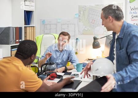 Middle aged Caucasian man discussing project with young Black man and middle aged Caucasian man in office, foreman gesturing while sitting at desk with construction plans visible Stock Photo