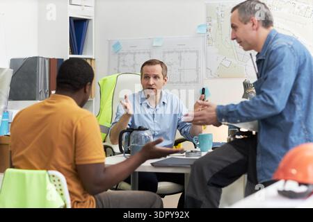 Middle aged Caucasian man sitting at desk gesturing while talking to middle aged Black man and middle aged Caucasian man, standing in office with architectural plans visible Stock Photo