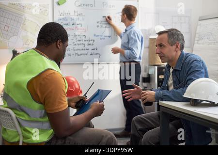 Middle aged Caucasian man discussing construction project details with young adult Black man holding tablet in office, while another middle aged Caucasian man writing on whiteboard in background Stock Photo