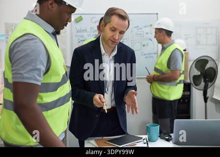 Middle aged Caucasian man in suit discussing project with young Black man in safety vest, while another middle aged Caucasian man in hard hat working in office with construction plans Stock Photo