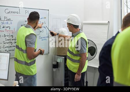 Two middle aged Caucasian men wearing safety vests and hard hats standing in office discussing construction project, one writing on whiteboard while other pointing at diagram Stock Photo