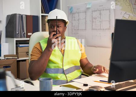 Middle aged Black man wearing safety vest and hard hat sitting at office desk, using walkie talkie and working on computer, with construction blueprints visible in background Stock Photo