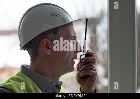 Middle aged Caucasian man wearing hard hat and safety vest standing in office using walkie talkie communicating instructions looking out window with focused expression Stock Photo