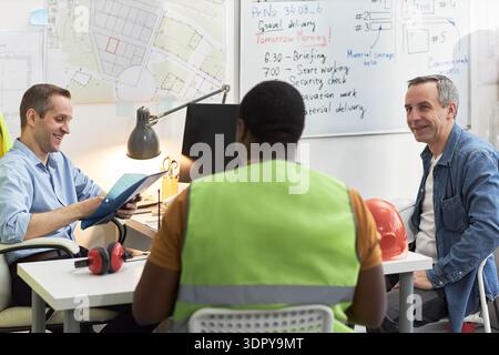 Middle aged Caucasian men and Black man discussing project details in office, foreman wearing safety vest sitting with colleagues near construction plans and safety helmet on table Stock Photo