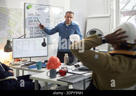 Middle aged Caucasian man standing in office holding hard hat, explaining project plan on whiteboard to multiethnic team of foremen sitting at desk with construction documents Stock Photo