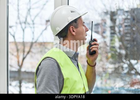 Middle aged Caucasian man wearing safety helmet and reflective vest standing in office holding walkie talkie, communicating instructions while looking out window Stock Photo