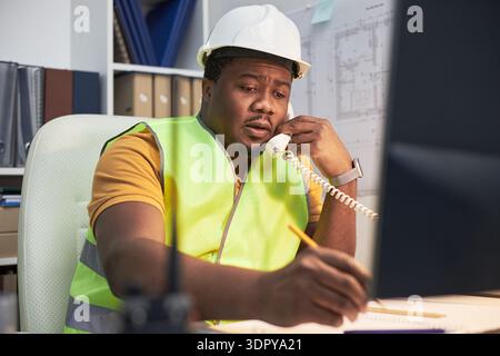 Black middle aged man wearing safety vest and hard hat sitting at office desk, talking on landline phone while writing notes and looking at computer monitor with construction plans visible Stock Photo