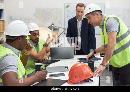 Middle aged Caucasian man in suit supervising group of young adult multiethnic men in safety vests and helmets, discussing construction plans in office setting with blueprints and laptops Stock Photo