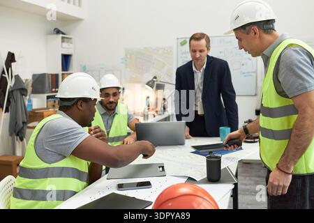 Multiethnic group of middle aged men wearing safety vests and hard hats discussing construction plans, with Caucasian middle aged foreman standing by desk in modern office Stock Photo