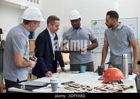 Group of middle aged men including Caucasian and Black foreman discussing construction plans in office, standing around table with blueprints and safety helmets, collaborating on project Stock Photo