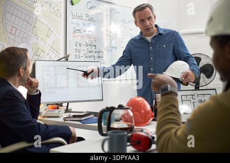 Middle aged Caucasian man standing in office holding hard hat and pointing at computer monitor while discussing construction plans with two colleagues during meeting Stock Photo