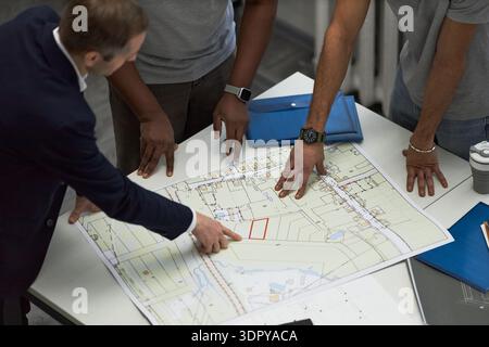 Middle aged Caucasian man and two young adult men, one Black and one possibly South Asian, reviewing architectural blueprint on office table, foreman pointing at building plan Stock Photo