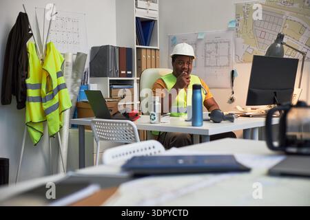 Black middle aged male construction site foreman wearing hard hat working at desk in office, using walkie talkie for communication, construction plans and safety vests visible in background Stock Photo