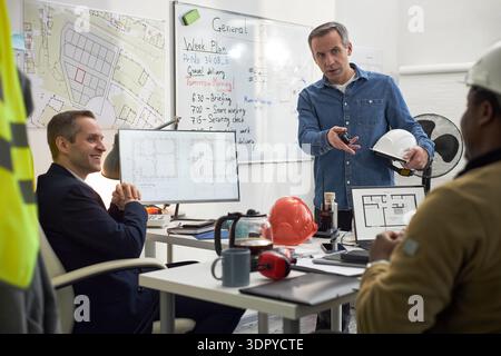 Middle aged Caucasian man standing and gesturing while holding hard hat, discussing construction plans with seated Black man and Caucasian man in office with architectural drawings visible Stock Photo