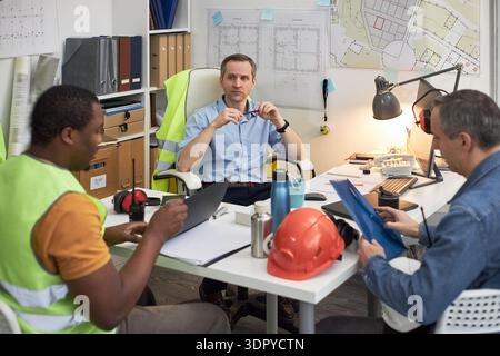 Caucasian middle aged man sitting at desk leading meeting with Black young adult man and Caucasian middle aged man in office, discussing construction plans and reviewing documents Stock Photo