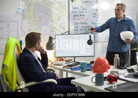 Middle aged Caucasian man standing and gesturing toward computer monitor with construction plans, while young adult Caucasian man sitting at desk listening in office with foreman context Stock Photo