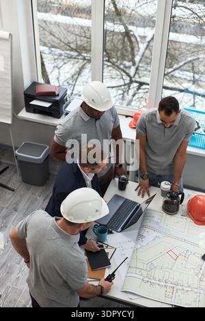 Vertical of group of middle aged men including Caucasian and Black foreman standing around office table discussing construction plans with laptop, blueprints, coffee pot and safety helmets visible Stock Photo