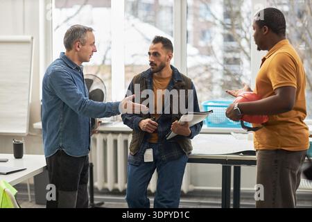Middle aged Caucasian man gesturing while talking to young adult Middle Eastern man and young adult Black man holding hard hat in office setting, discussing construction project Stock Photo
