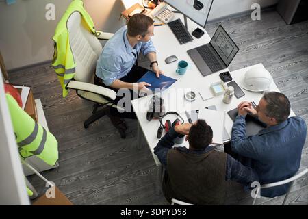 Three middle aged men including Caucasian foreman discussing construction project at office desk, reviewing blueprints and digital plans on laptop, safety vest and helmet visible nearby Stock Photo