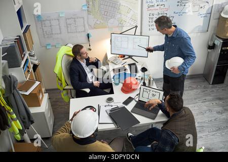 Group of middle aged Caucasian men and one Black man discussing construction blueprints in office, foreman standing and pointing at monitor while others sitting with laptops and hard hats Stock Photo