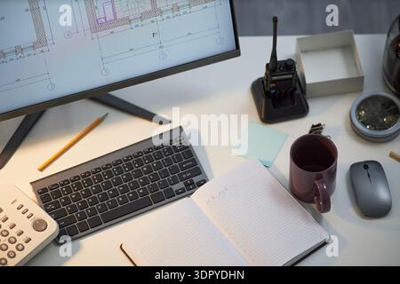Office desk showing open notebook, wireless keyboard, computer monitor displaying architectural plans, walkie talkie, calculator, pencil, coffee mug, mouse, compass, sticky notes Stock Photo