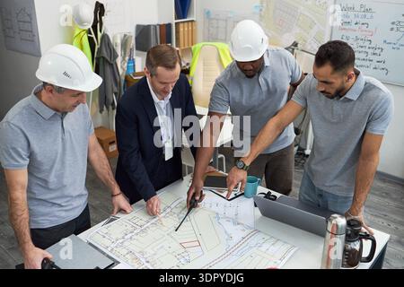 Group of middle aged men including Caucasian and Black foremen discussing blueprints in office, pointing at construction plans on table, collaborating on project management tasks Stock Photo